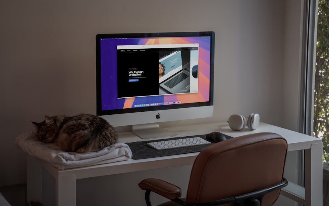A peaceful home office setup featuring an iMac showcasing a modern web design project, with a comfortable brown leather chair, wireless headphones, and a sleeping cat on a soft blanket. The natural light filtering through the window adds a calming atmosphere, making this an ideal space for creativity and productivity. Perfect inspiration for remote workers, designers, or anyone who loves minimalist workspaces with a touch of coziness.
